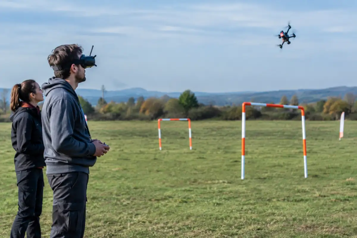 Un pilote en lunettes FPV dirige un quadricoptère en figure acrobatique sur un terrain dégagé, avec un observateur à proximité.