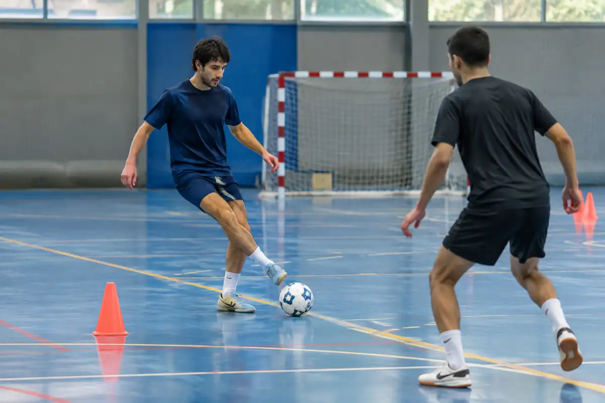 Deux joueurs travaillent la passe courte et le contrôle orienté au futsal dans une salle équipée de cônes et d’un but.