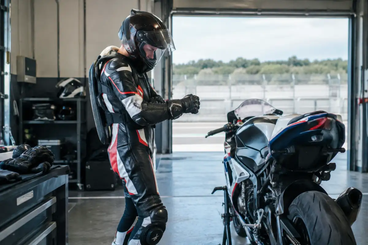 Pilote débutant vérifiant ses gants et sa combinaison en cuir près d’une moto de circuit, avec casque intégral et bottes