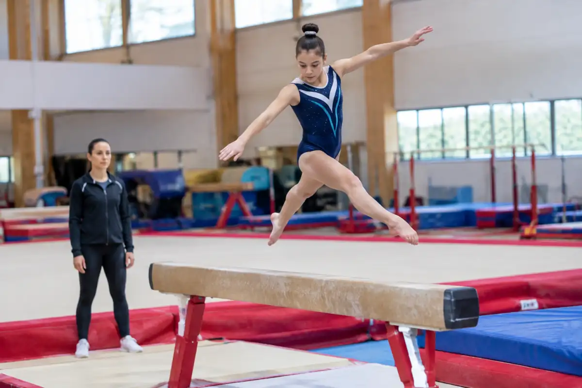 Gymnastique artistique féminine : une gymnaste réalise un élément acrobatique sur une poutre dans une salle équipée de tapis.