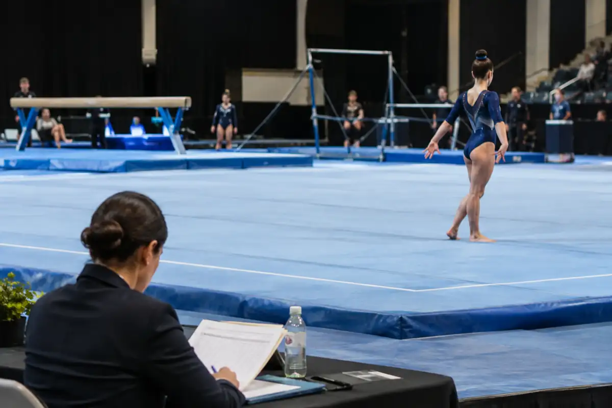 Une juge consulte sa feuille de notes après une réception au sol, avec les agrès de gymnastique artistique visibles en arrière-plan.