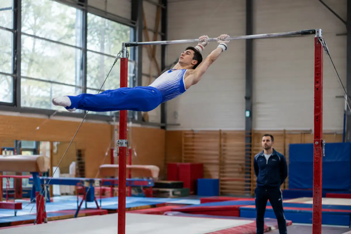 Gymnaste de gymnastique artistique masculine en plein balancement à la barre fixe, dans une salle équipée de tapis.