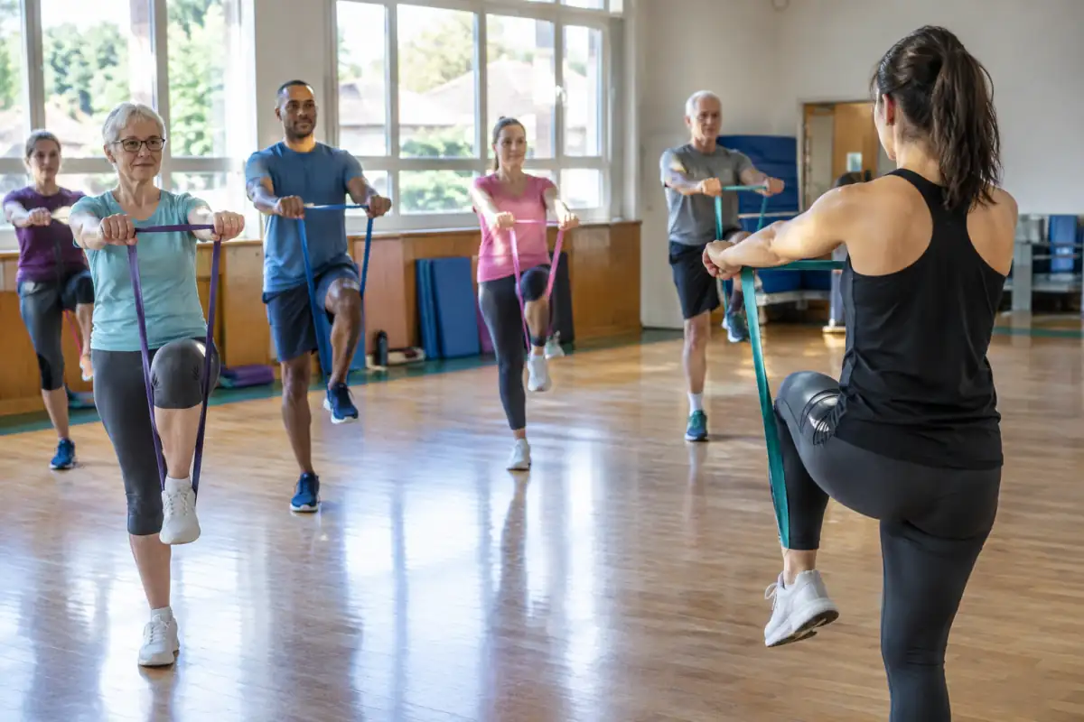 Un groupe mixte suit un cours de gymnastique volontaire en salle, avec des exercices d’équilibre et un élastique de résistance.