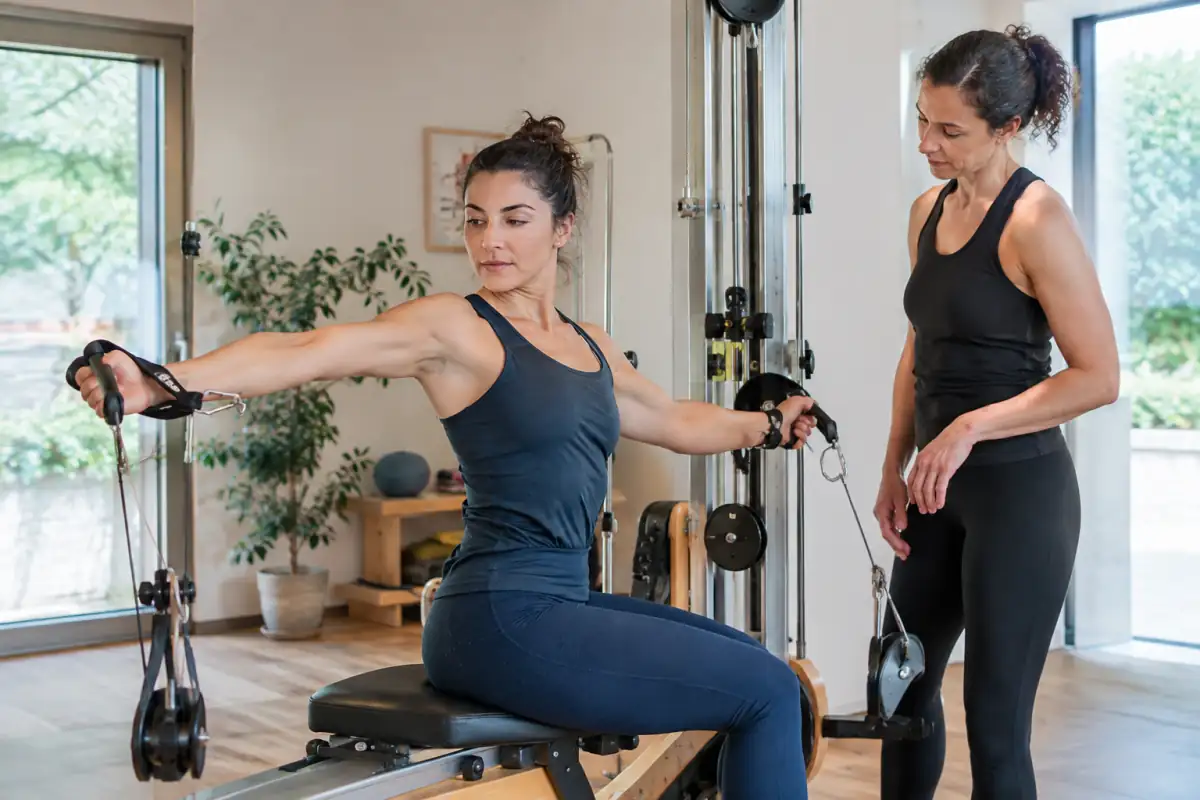 Une pratiquante réalise un mouvement circulaire sur un appareil à poulies de Gyrotonic, guidée par un enseignant en studio.
