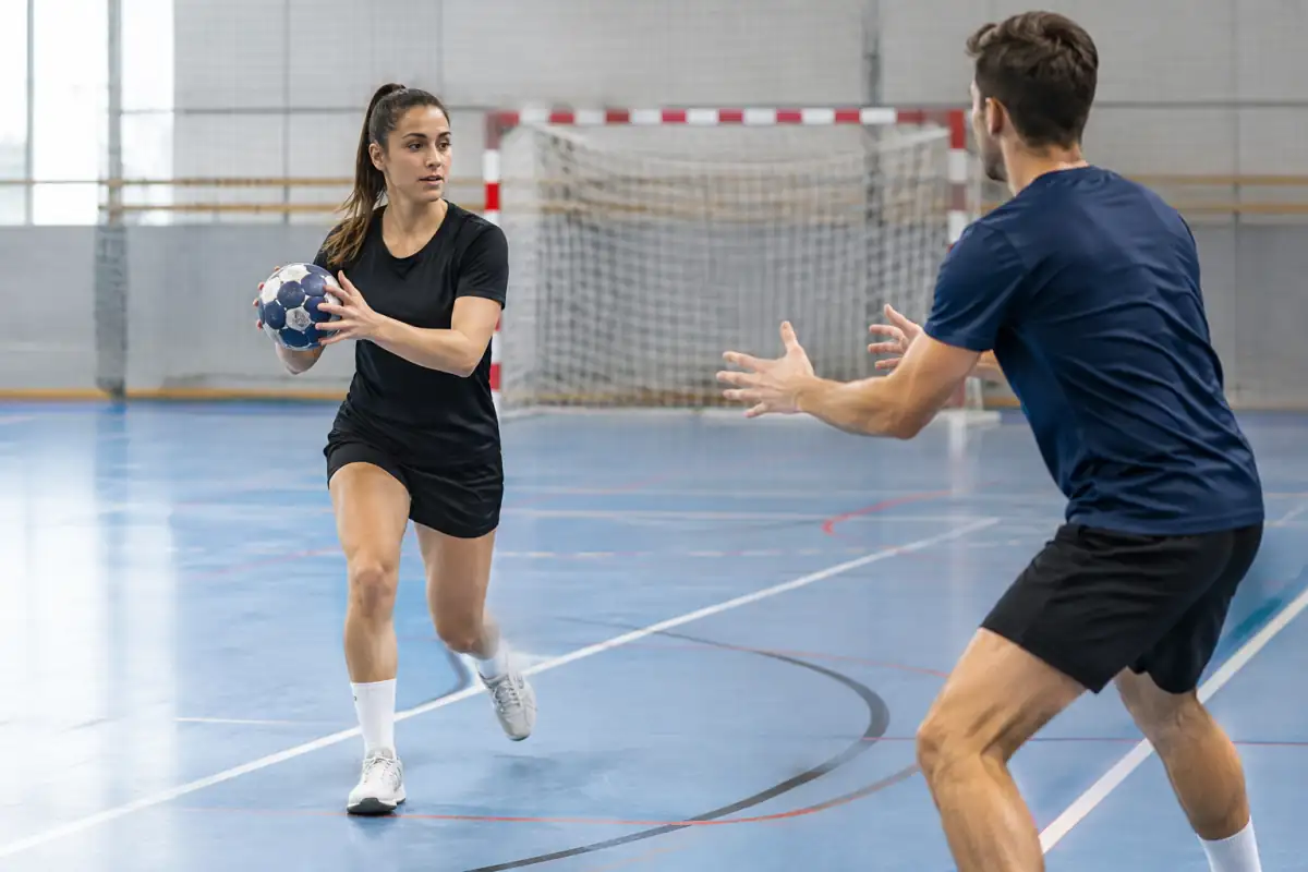 Deux débutants travaillent une passe et une réception en mouvement sur un terrain de handball en salle.
