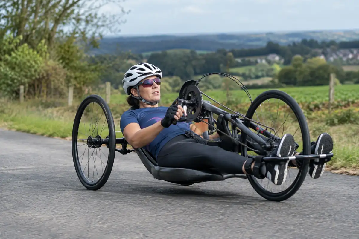 Athlète en handbike couché actionnant les manivelles sur une route de campagne, avec casque et gants.