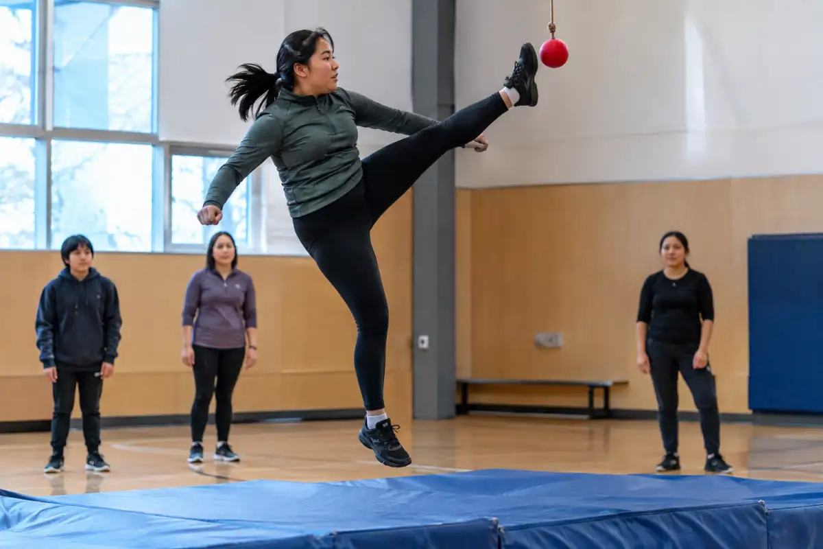 Un athlète réalise un high kick inuit en sautant sur un pied pour toucher une balle souple suspendue dans une salle sportive.