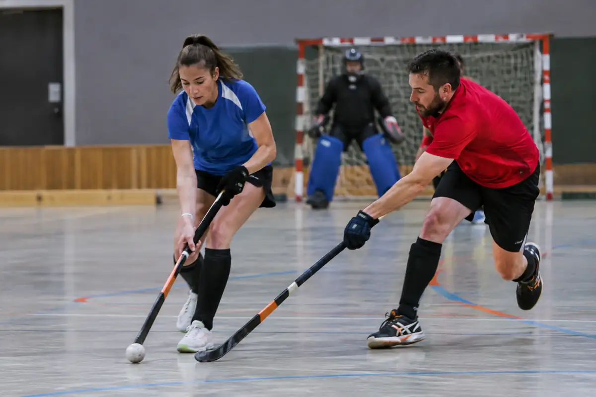 Match de hockey en salle dans un gymnase : deux joueurs disputent la balle près des bandes, face au gardien.