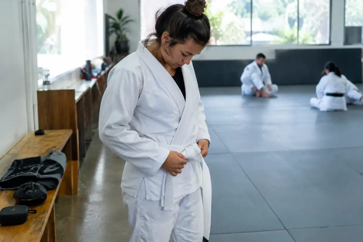 Débutant en jiu-jitsu brésilien ajustant la ceinture de son kimono avant un entraînement, avec rashguard et short posés près du tatami.