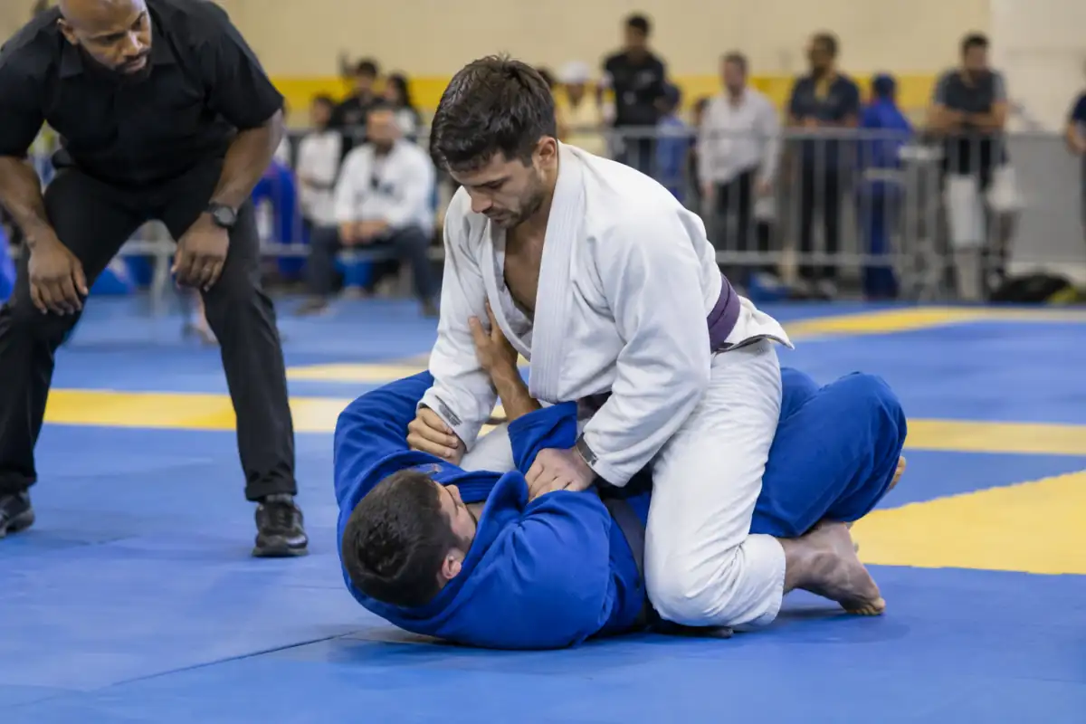 Combat de jiu-jitsu brésilien en gi : un compétiteur contrôle un genou sur le ventre sous le regard attentif de l’arbitre.