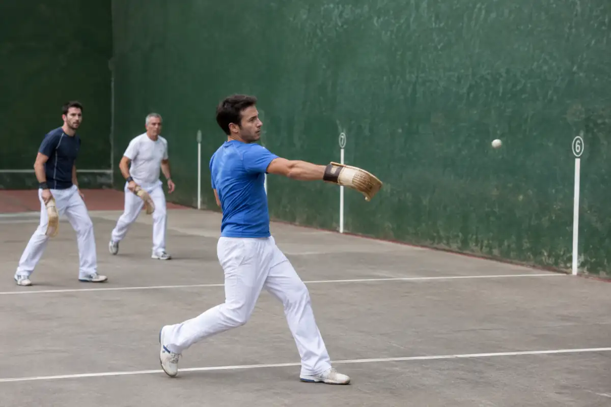 Joueur de joko garbi frappant une pelote avec une petite chistera sur un fronton basque en plein air.