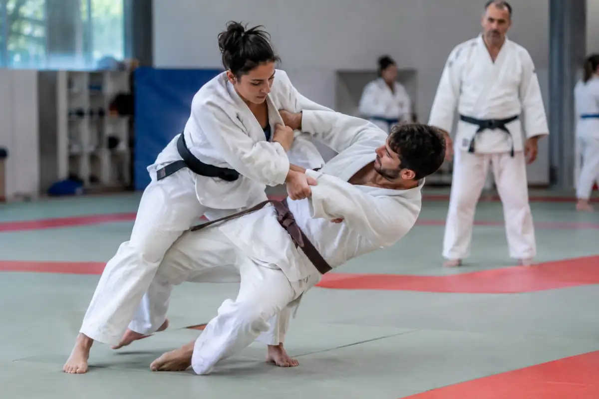 Deux judokas en judogi travaillent une projection debout et une chute contrôlée sur le tatami d’un dojo.