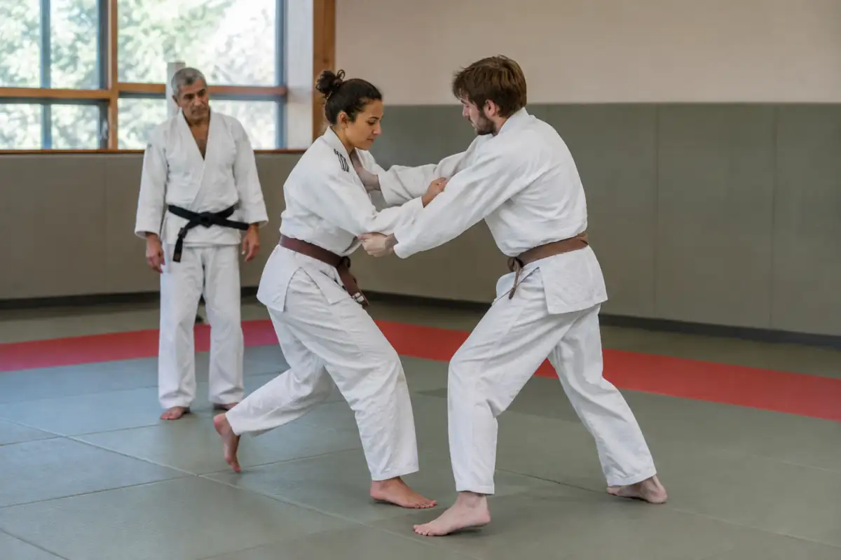 Deux débutants en judogi répètent lentement une entrée de projection sur tatami, sous la supervision attentive de leur professeur.