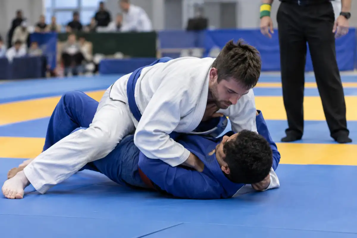Deux compétiteurs en kimono travaillent un contrôle au sol en Ne-Waza sous la surveillance d’un arbitre lors d’une rencontre de jujitsu.