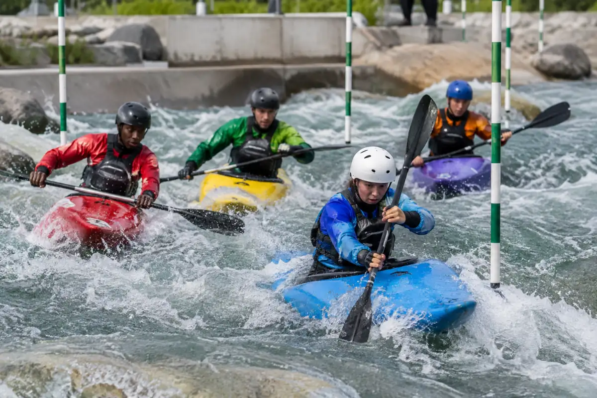 Quatre kayakistes équipés de casques et de gilets se disputent une porte sur un parcours de kayak cross en eau vive.