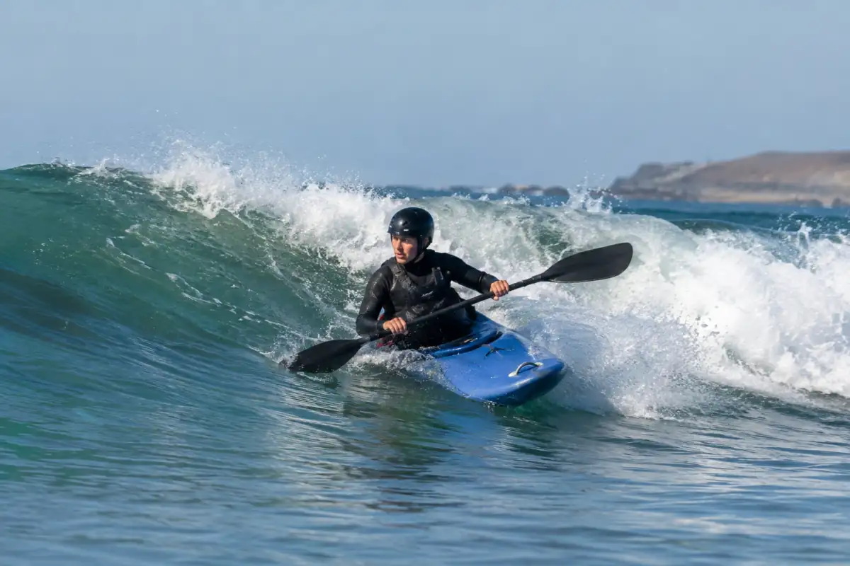 Un kayakiste en combinaison et casque trace un virage à la pagaie sur la face d’une vague en kayak surf.