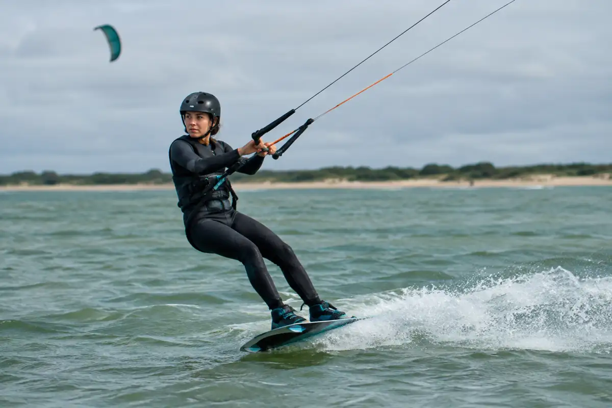 Un kitesurfeur en combinaison glisse sur une planche twin-tip, tracté par une aile au-dessus de l’eau.