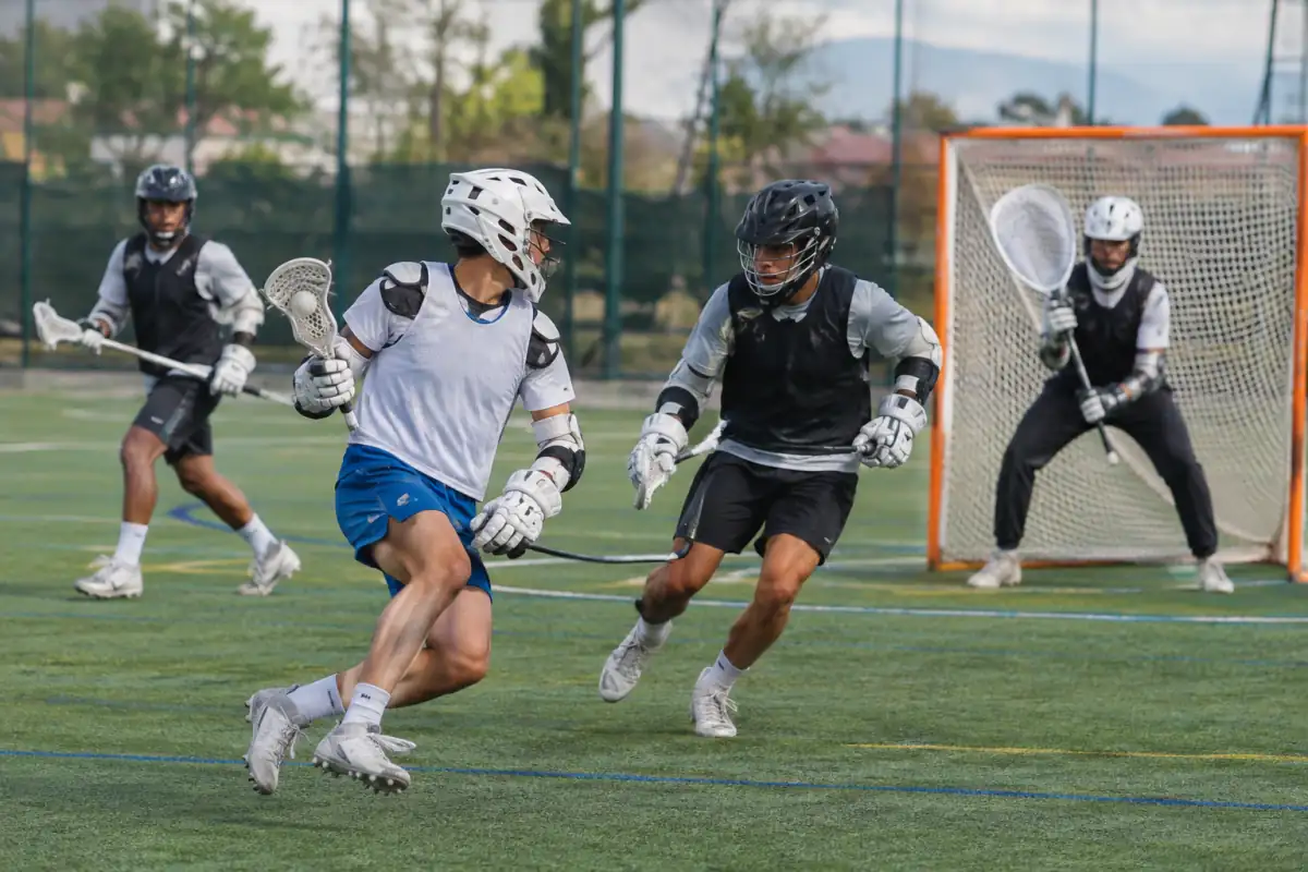 Un joueur attaque le but en lacrosse sixes avec sa crosse, tandis qu’un défenseur et le gardien protègent la zone.