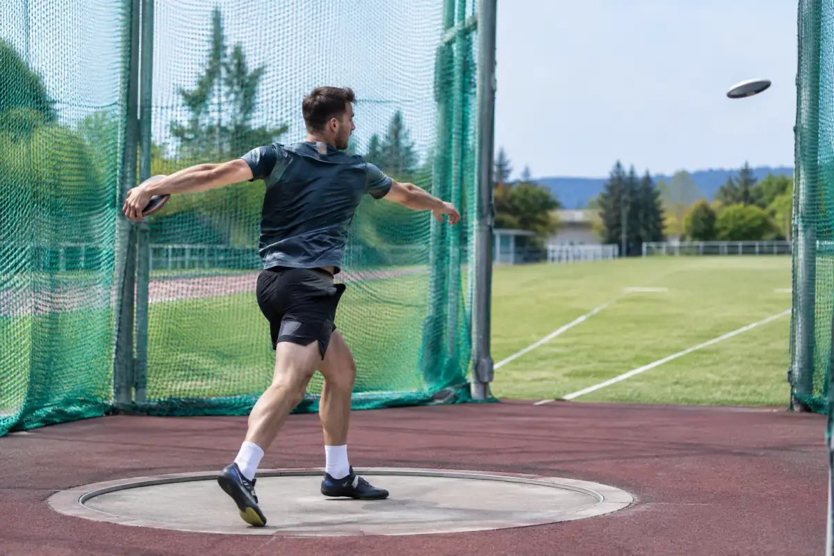Athlète en rotation dans le cercle lors d’un lancer du disque sur un stade d’athlétisme équipé d’une cage de protection.