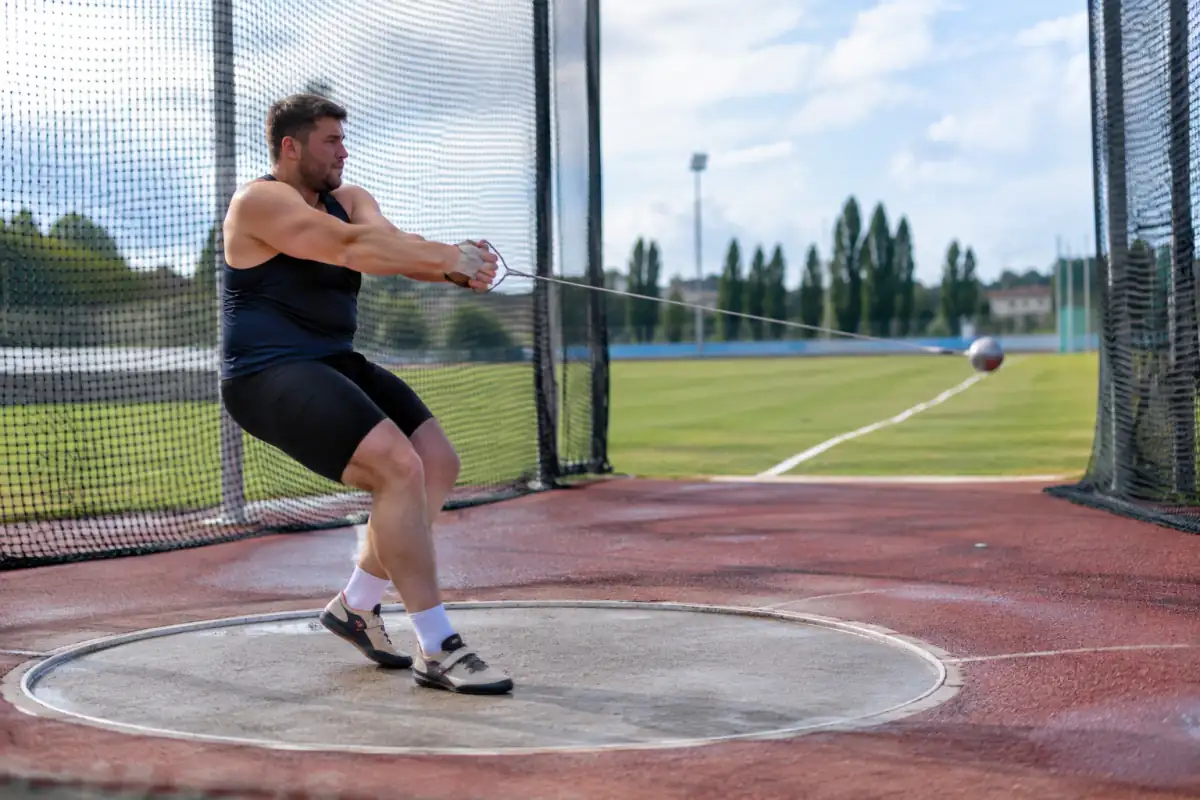 Un athlète effectue une rotation au lancer du marteau dans un cercle entouré d’une cage de protection sur un stade.