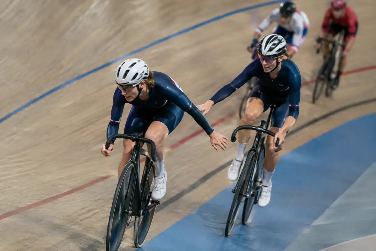 Relais à la main entre deux équipiers en Madison, sur des vélos de piste dans un vélodrome couvert.