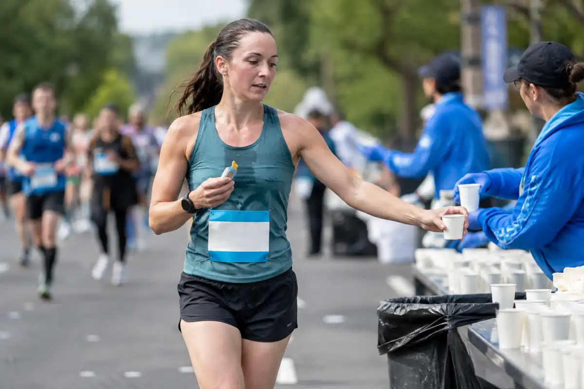 Un marathonien prend un gel énergétique en approchant d’un poste de ravitaillement, avec des gobelets d’eau et d’autres coureurs en course.