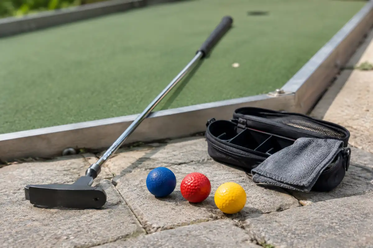 Putter, balles spécialisées aux finitions variées et sac compartimenté posés près d’une piste de mini-golf de club.