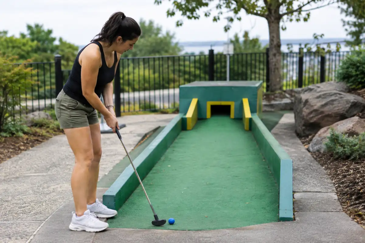 Joueur de mini-golf en posture stable, alignant son putter et sa balle vers l’ouverture étroite d’une piste à obstacles.
