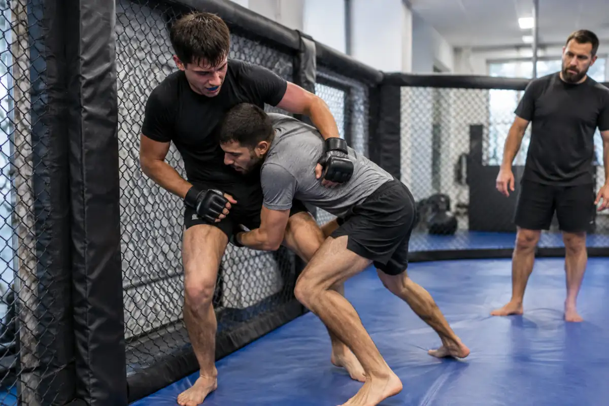 Deux pratiquants travaillent une amenée au sol en MMA dans une cage d’entraînement, sous le regard de leur entraîneur.