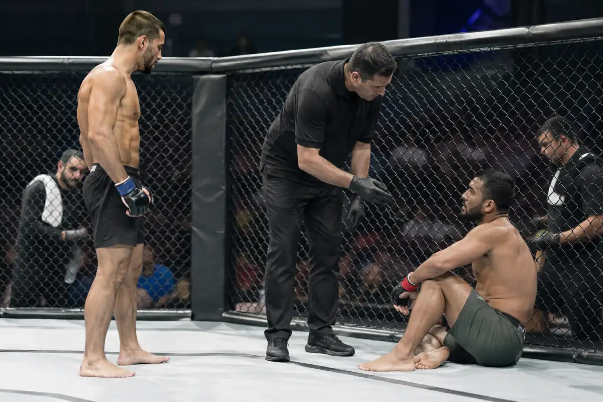 Un arbitre vérifie l’état de deux combattants dans une cage de MMA pendant la pause entre deux rounds.