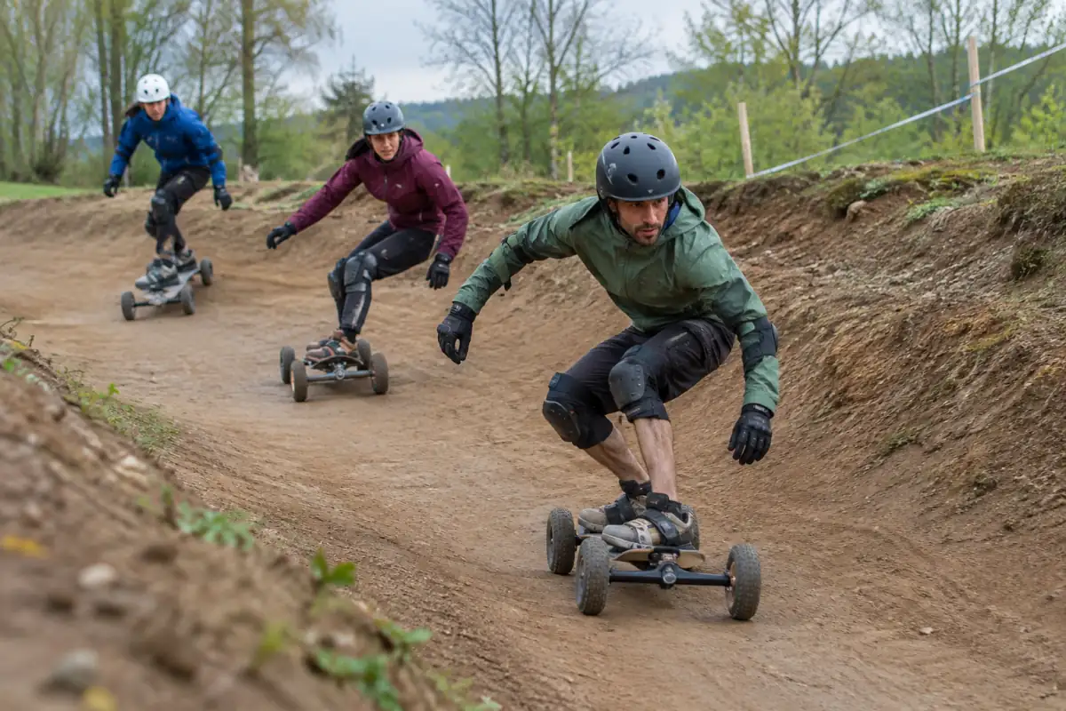 Des riders équipés négocient un virage en terre lors d’une course de mountainboard cross sur une piste tout-terrain.