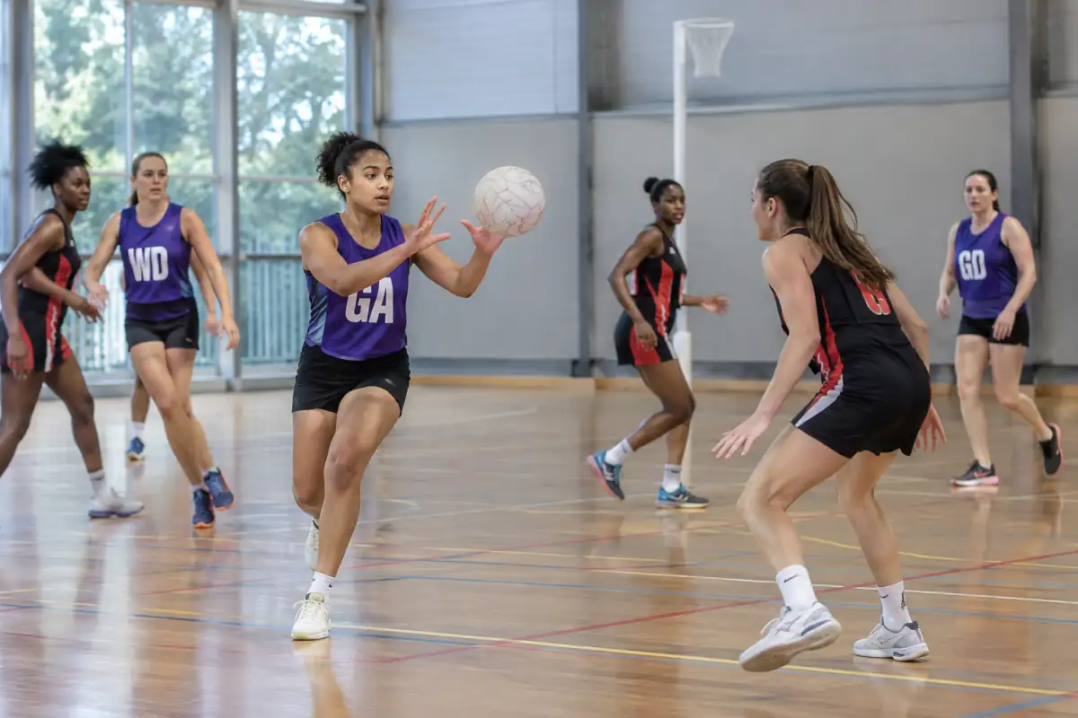 Une joueuse de netball reçoit le ballon sur un terrain intérieur, sous la défense d’une adversaire à distance réglementaire.