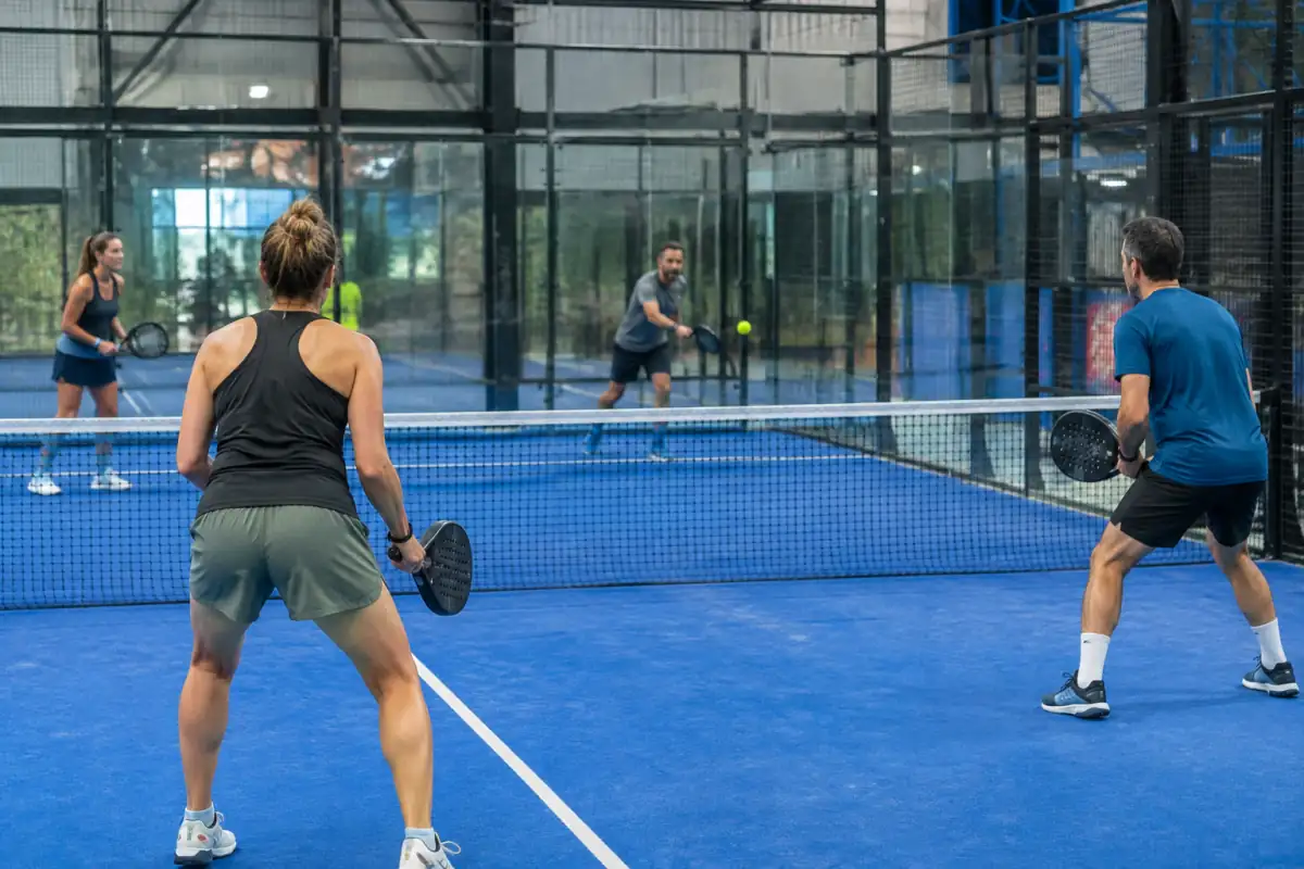 Échange de padel en double sur un court vitré, un joueur renvoie la balle après son rebond contre la paroi.