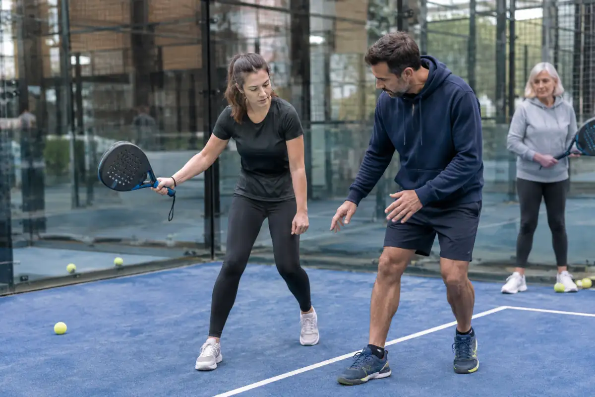 Deux débutants découvrent le coup droit au padel avec un encadrant, une raquette à dragonne et une vitre en fond de court.