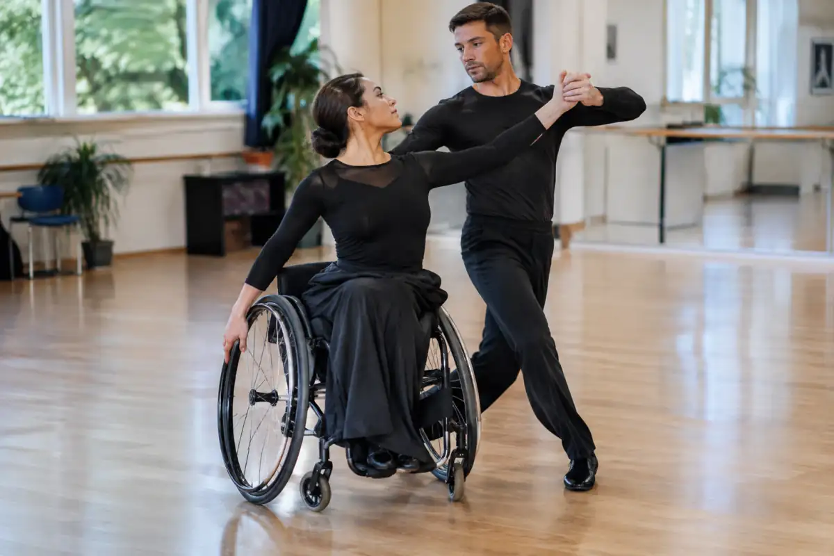 Couple de para-danse sportive en action : danseuse en fauteuil manuel et partenaire debout synchronisent une figure en salle.