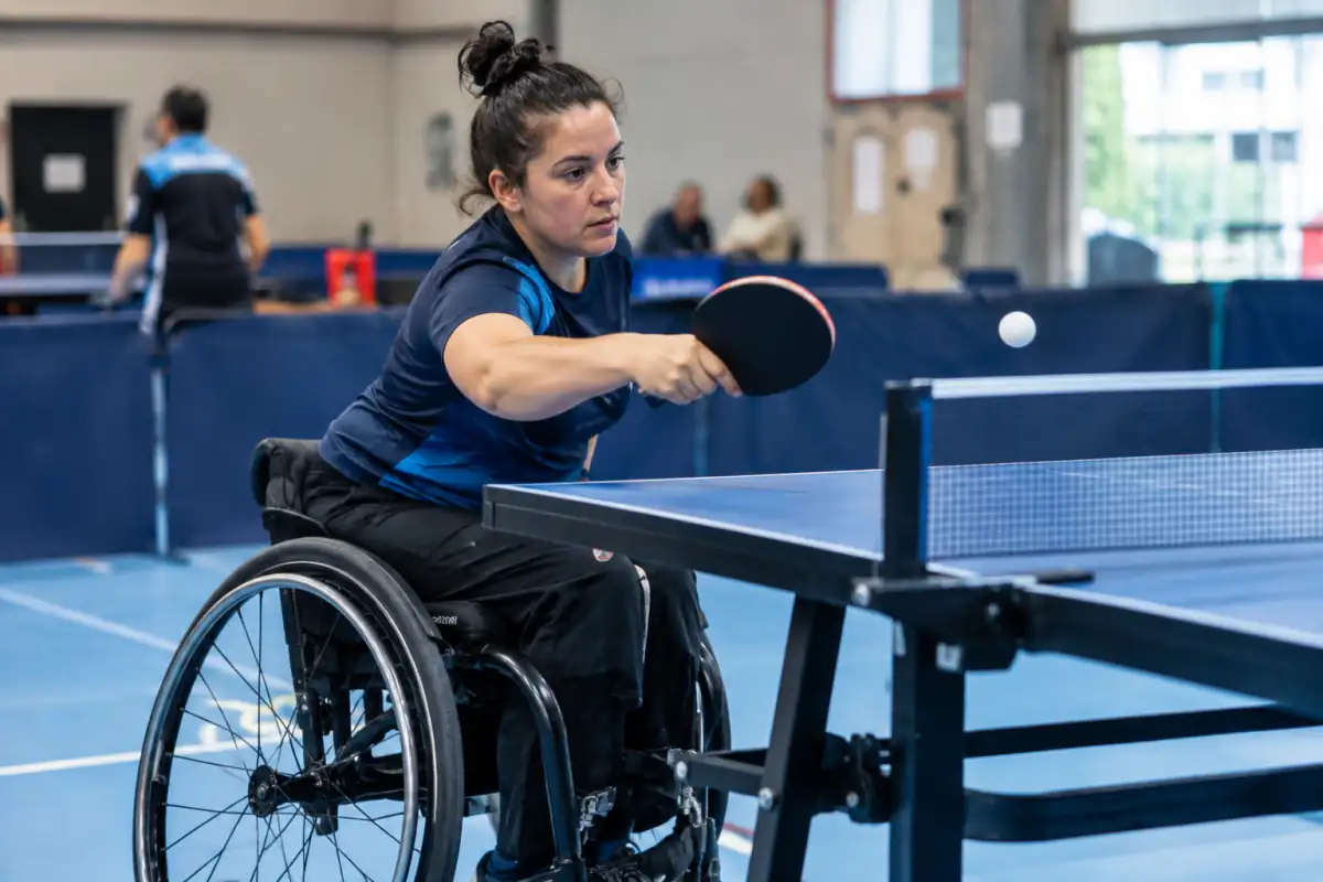 Joueur de para-tennis de table en fauteuil roulant effectuant un coup droit près de la table pendant un échange en salle.