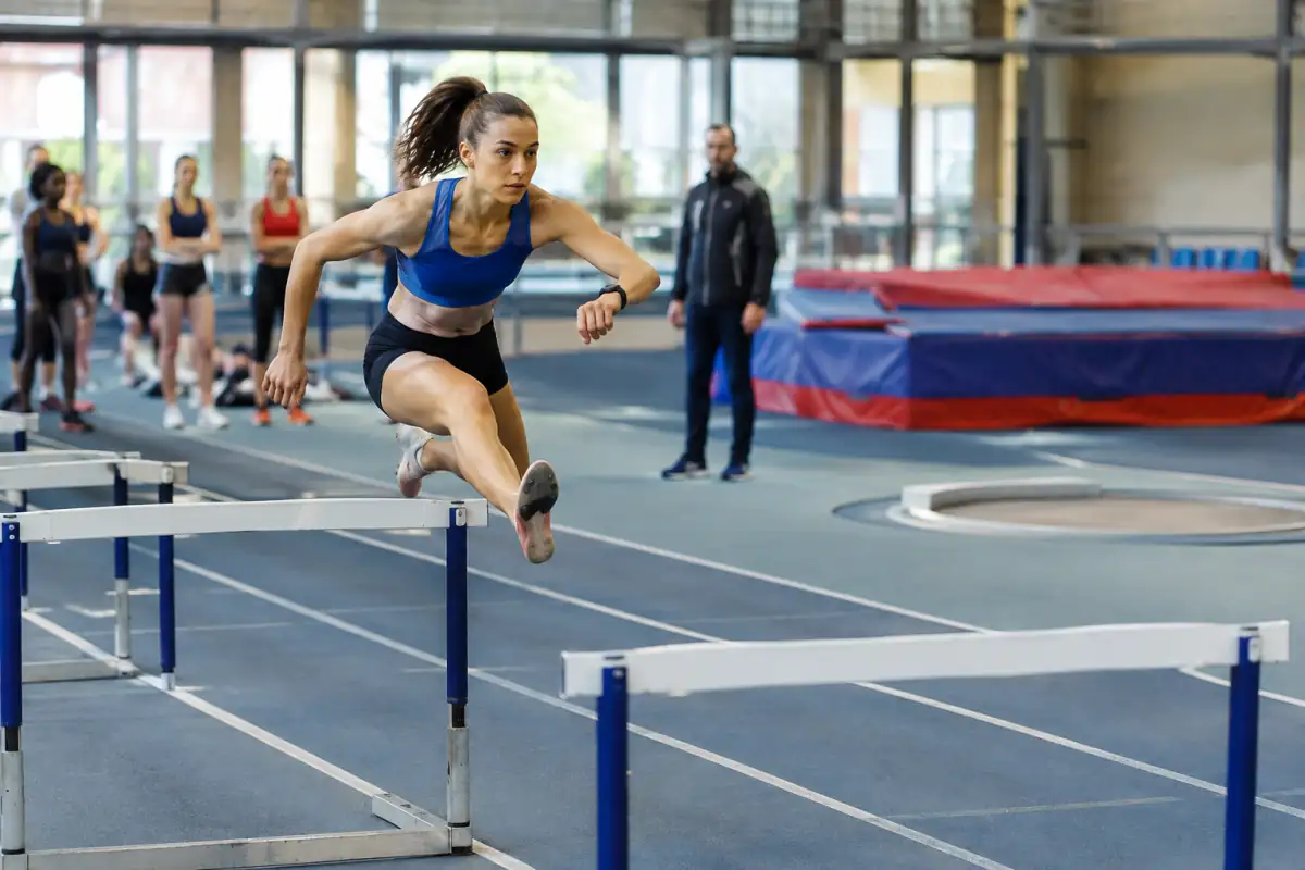 Athlète franchissant une haie lors d’un pentathlon en salle sur une piste d’athlétisme couverte.