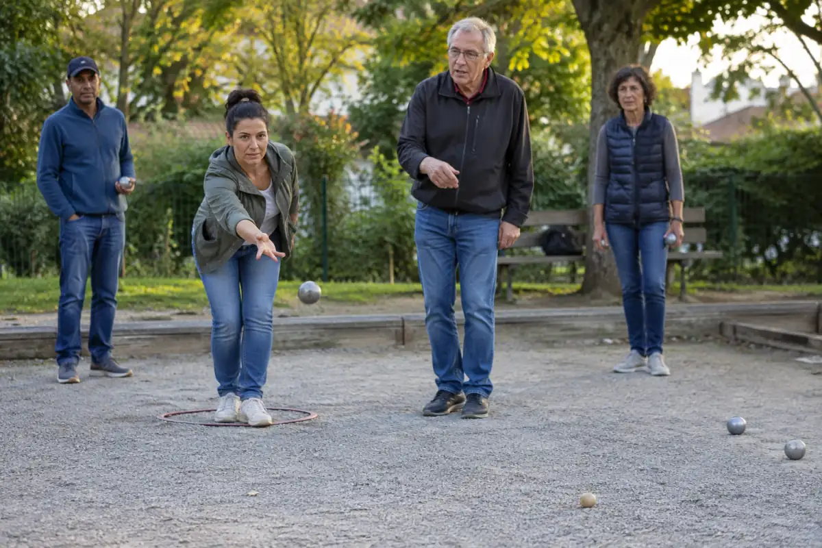 Un débutant lance une boule sur un terrain de pétanque tandis qu’un partenaire lui explique le jeu à distance.