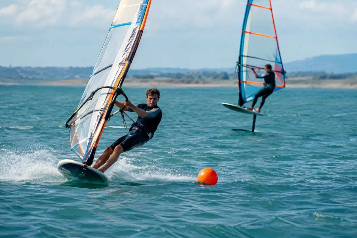 Un véliplanchiste négocie un jibe de slalom près d’une bouée, tandis qu’un autre navigue en foil au large.