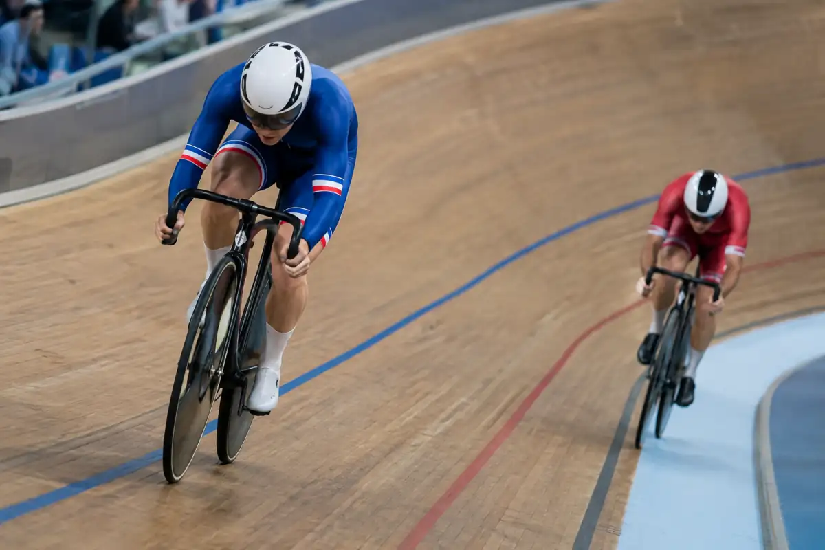 Deux cyclistes en poursuite individuelle sur piste pédalent à pleine vitesse sur un vélodrome couvert.