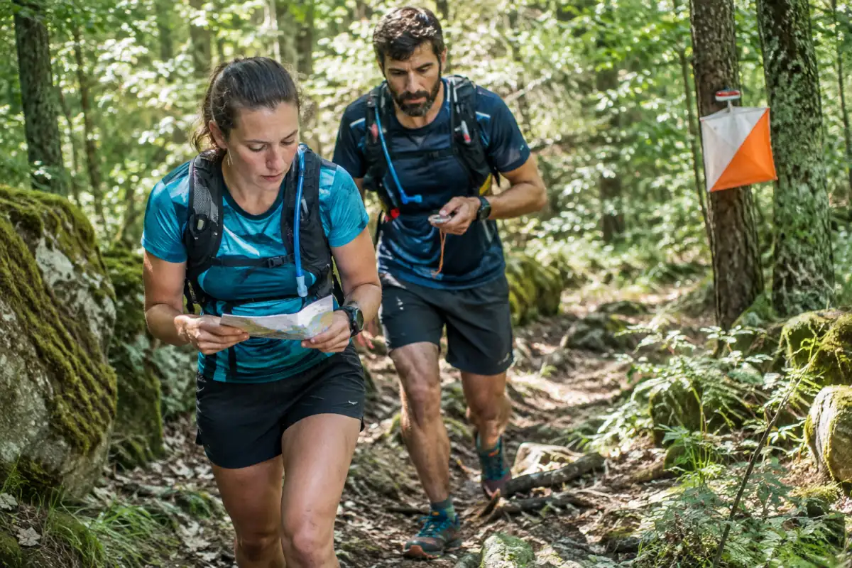 Équipiers en raid multisport nature consultent une carte et une boussole sur un sentier forestier, sacs d’hydratation au dos.