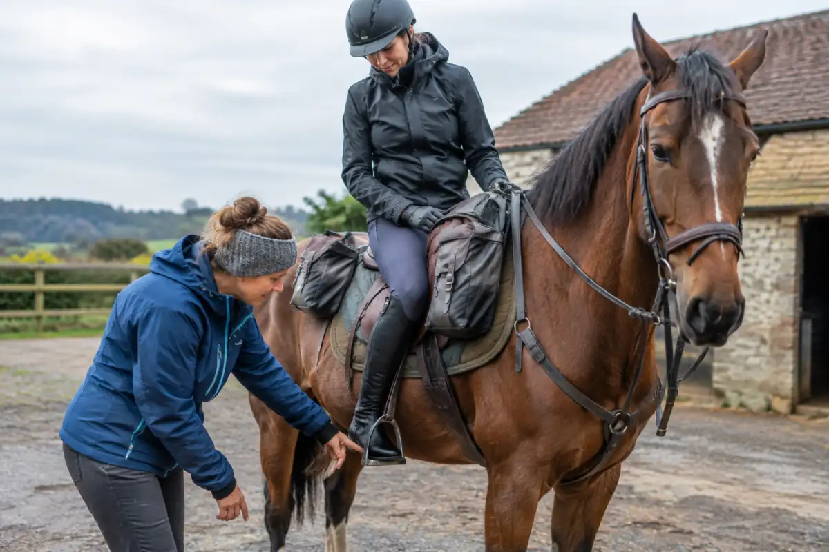 Cavalière équipée d’un casque ajusté, d’un pantalon d’équitation et de chaussures à talon, près d’un cheval sellé avant une randonnée.