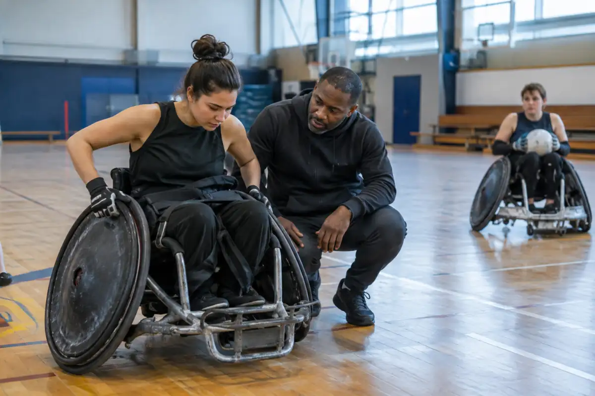 Un débutant en fauteuil de rugby travaille le freinage dans un gymnase, accompagné par un entraîneur qui vérifie son installation.