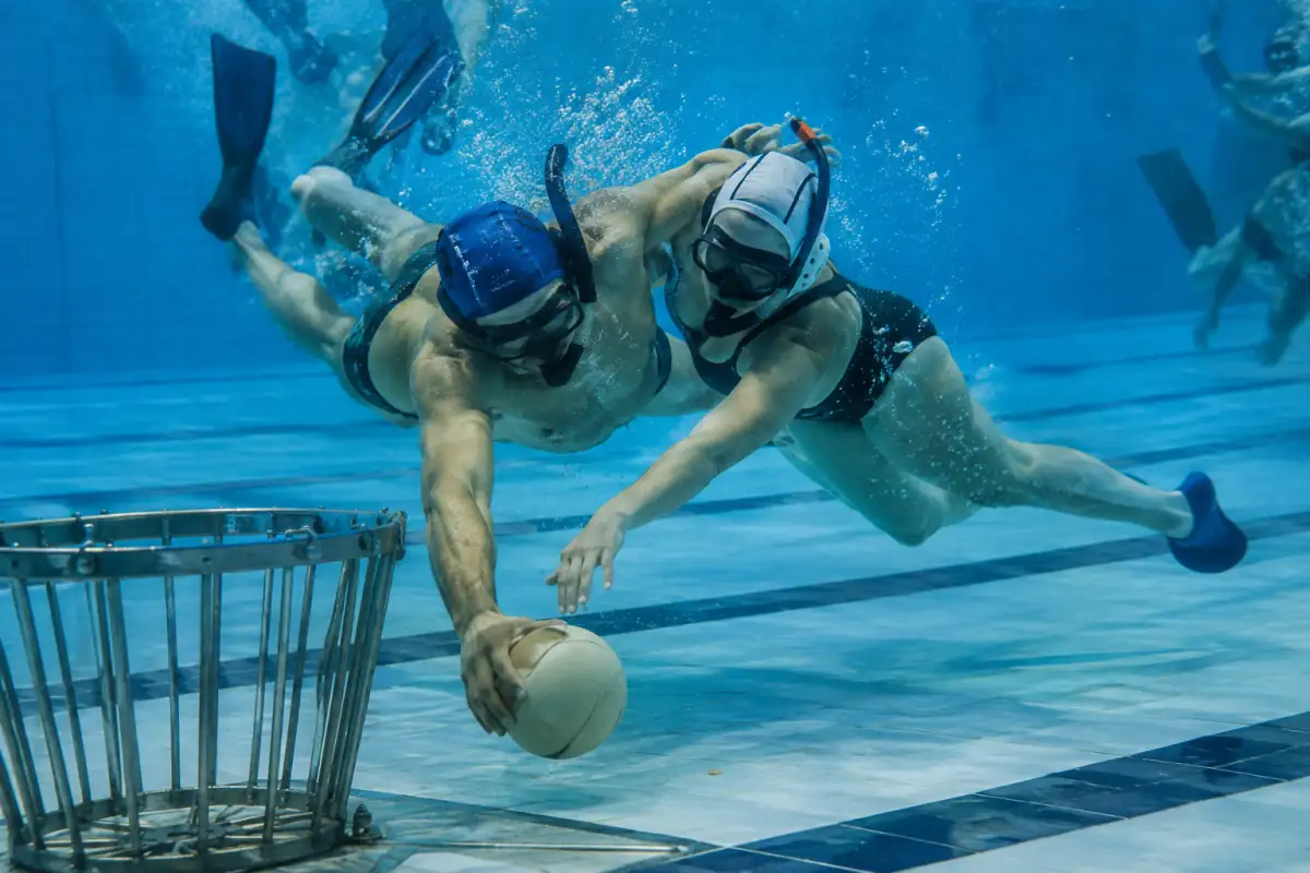 Deux joueurs disputent un ballon lesté près du panier au fond d’une piscine lors d’un match de rugby subaquatique.