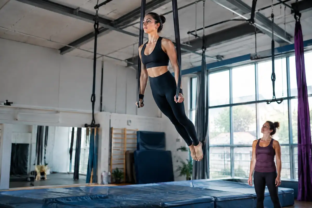 Artiste en suspension sur deux sangles aériennes dans une salle de cirque équipée de matelas de réception.