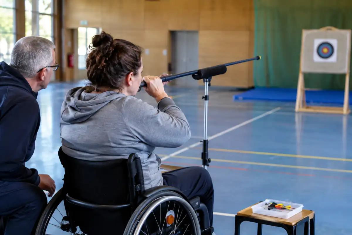 Une personne en fauteuil utilise une sarbacane adaptée sur un support réglable pour viser une cible dans une salle de sport.