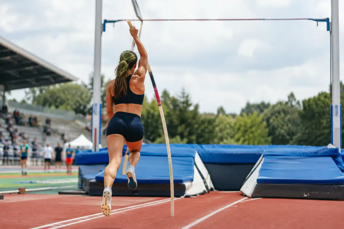 Athlète en plein saut à la perche, plantant une perche flexible sur une piste d’athlétisme devant la barre et le matelas.