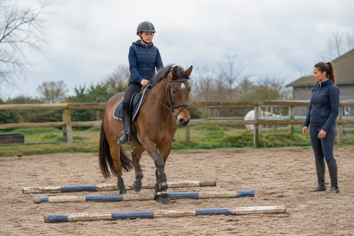 Un cavalier débutant franchit des barres au sol au trot dans une carrière, sous le regard de son enseignante de CSO.