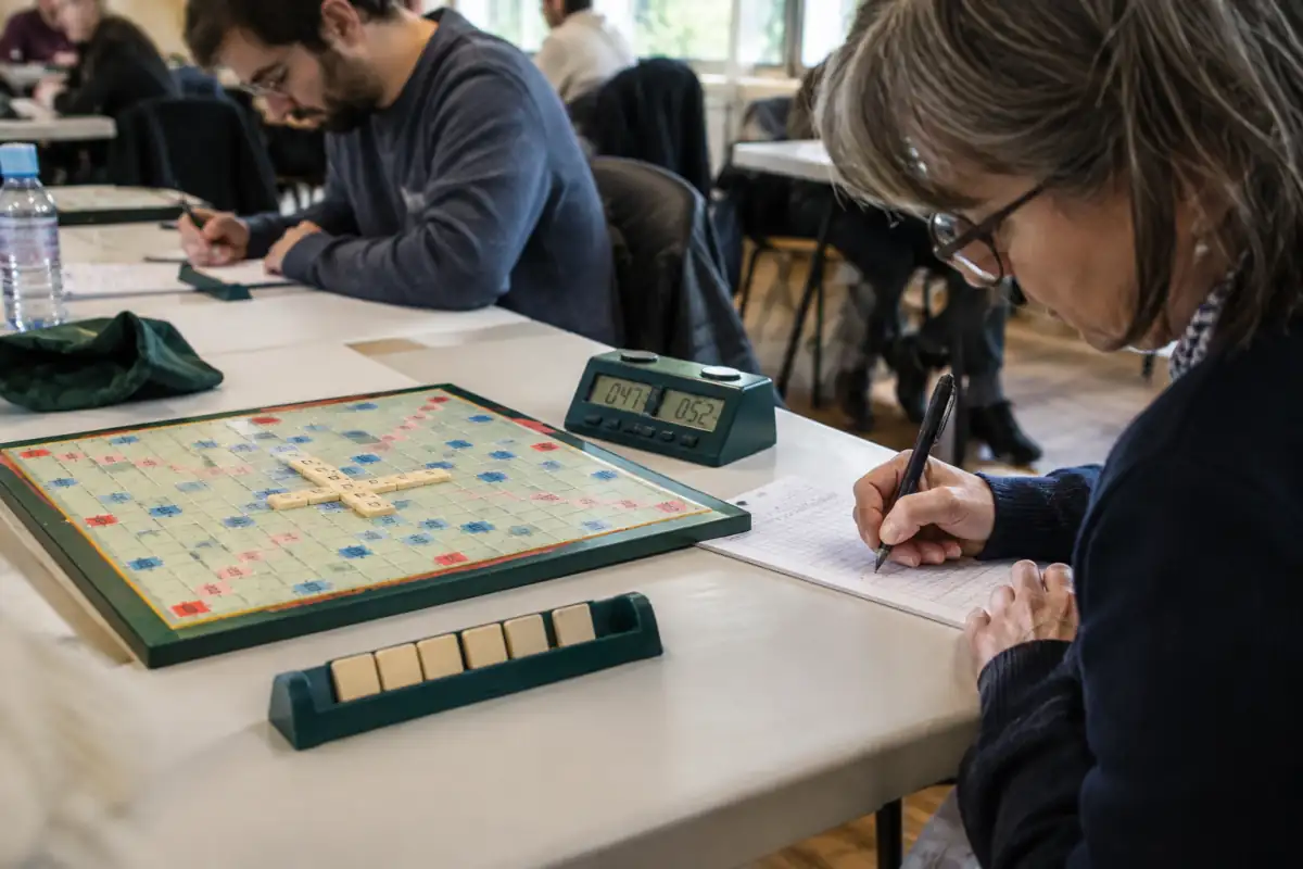 Joueur de Scrabble duplicate notant son mot et ses coordonnées devant la grille commune et son tirage de sept lettres.