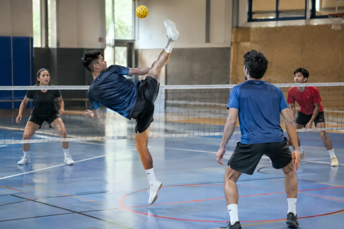 Joueurs de sepak takraw disputant un échange au-dessus du filet avec une balle tressée dans un gymnase.